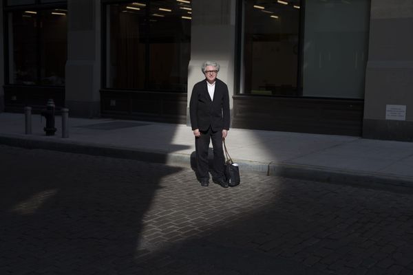 Boris Groys in a black suit stands alone on a city sidewalk in a spotlight of sunlight, holding a black bag, with a building and street in the background.