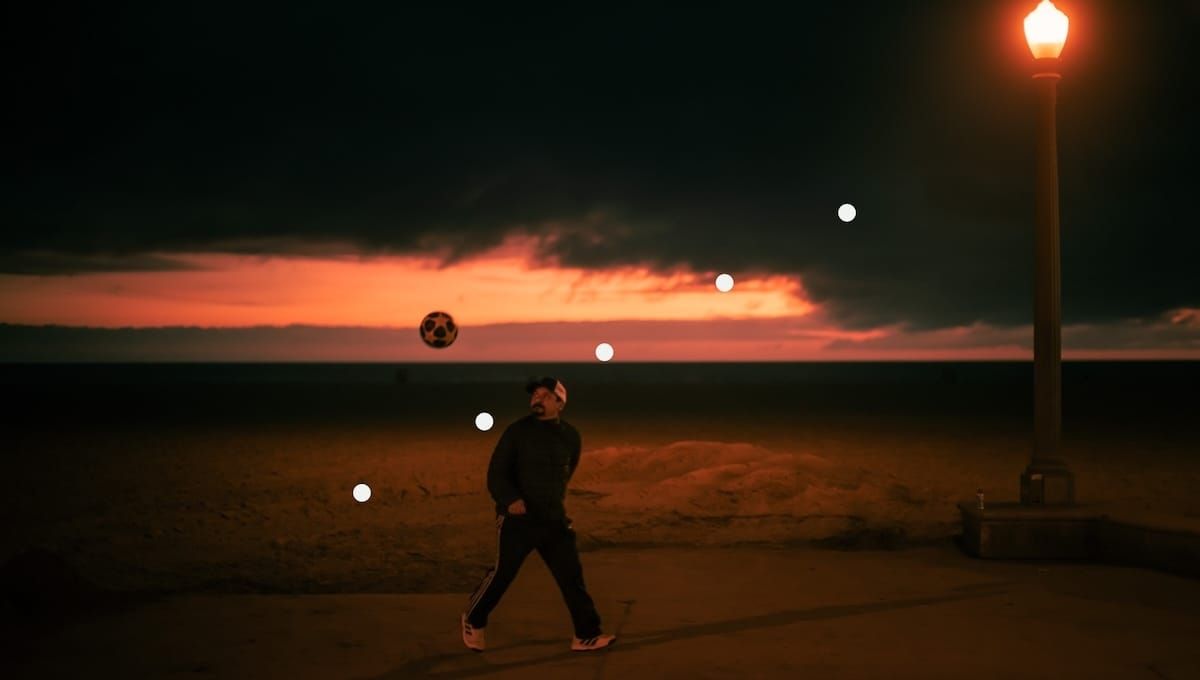 Photo of a man with a soccer ball mid-air, at the beach, at sunset.