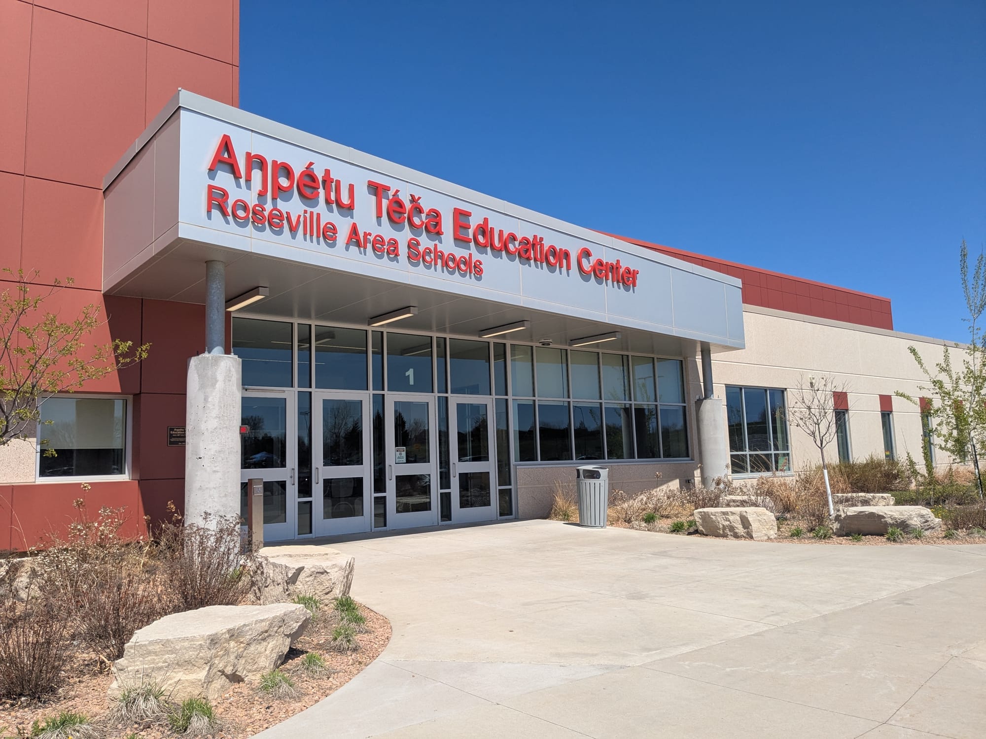 The main entrance to Aŋpétu Téča Education Center has glass windows and doors, surrounded by gray metal trim. The name of the center is written above in red metal lettering. 