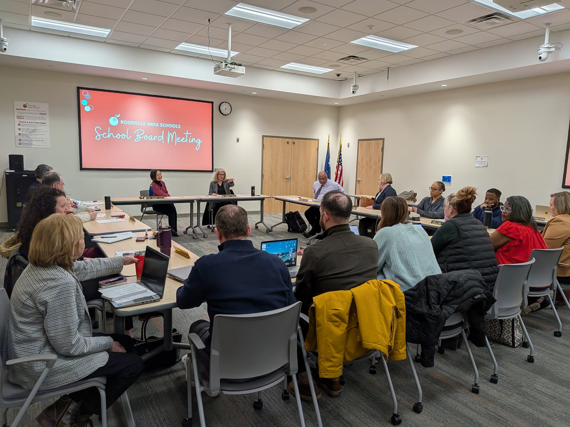 Sixteen adults sitting around tables arranged in a square. At the front and center facing the camera are School Board Chair Rose Chu and Superintendent Jenny Loeck