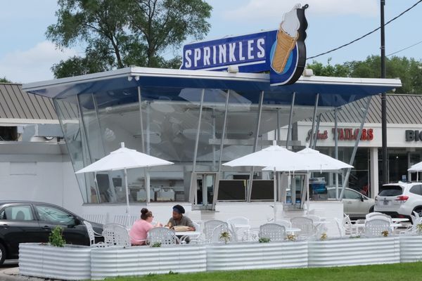 The retro blue and white building has angled glass windows and a blue and white sign on top that says Sprinkles next to an image of an ice cream cone. Two people are sitting at a white table.