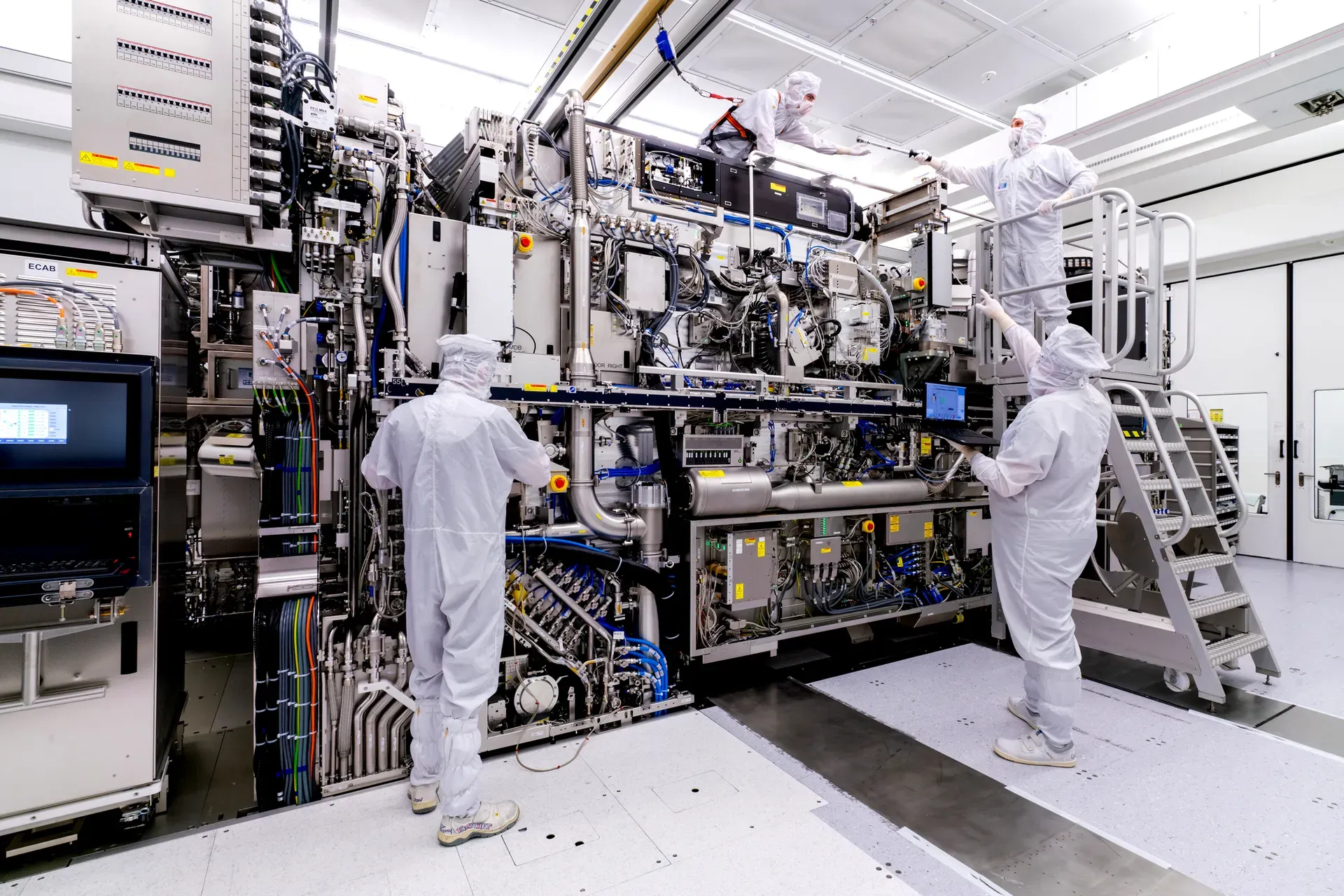 Three ASML technicians in white cleanroom suits and protective gear work on an EUV lithography system in a bright, sterile facility. One technician stands at floor level at the front of the machine, a second works at the right side using a metal access staircase, and a third is positioned at the top of the machine. The system itself is a massive wall of interlocking components including cables, pipes, conduits, and electronic modules in silver, blue, and yellow. A computer monitor is visible to the left. The machine fills nearly the entire frame.