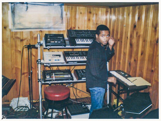 A young man stands in a wood-paneled home studio, surrounded by a rack of vintage synthesizers, drum machines, and sequencers, with a keyboard in front of him and a red stool nearby.