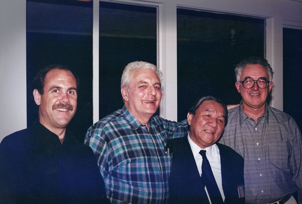 Four men, the synthesizer pioneers Dave Smith, Bob Moog, Ikutaro Kakehashi, and Tom Oberheim, posing together and smiling indoors at night.