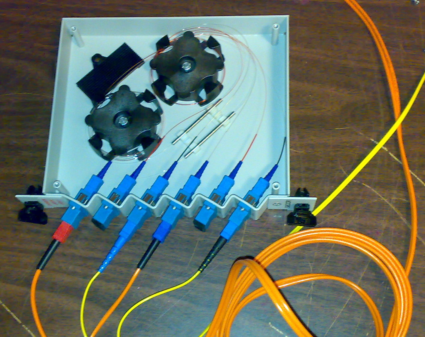 A fiber optic patch panel with six blue SC connectors mounted in a splice tray, with coiled orange and yellow fiber cables extending from the panel across a wooden surface.