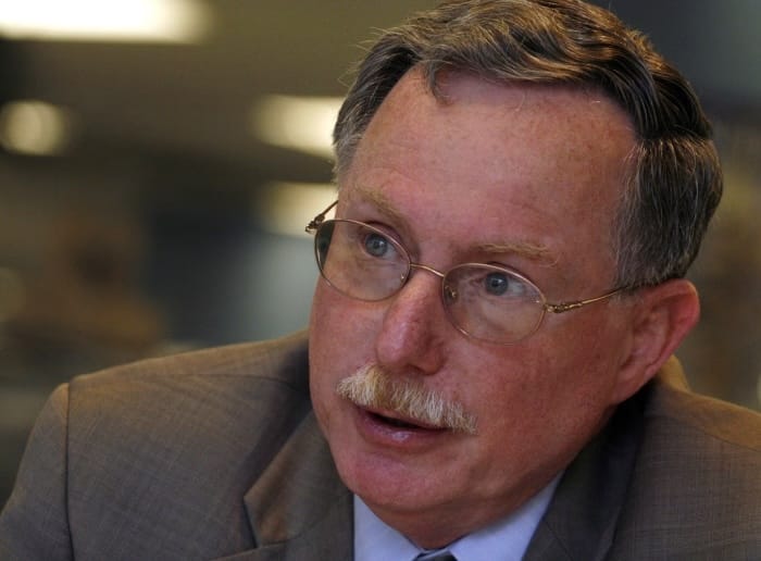 Close-up portrait of Mark Klein, an older man with gray hair, a gray mustache, and wire-rimmed glasses, wearing a gray suit, speaking mid-conversation with a blurred indoor background.