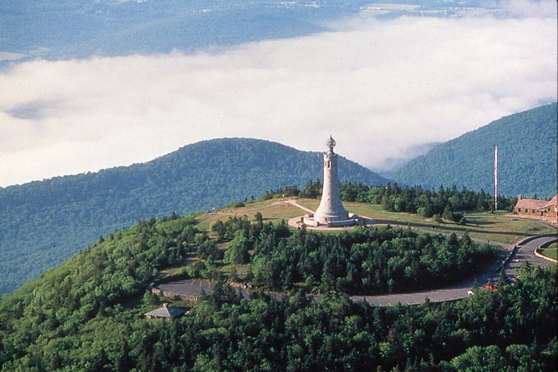 Aerial view of Mount Greylock's summit, showing the stone War Memorial Tower surrounded by forested peaks with low clouds in the valley, plus a distant radio tower and stone lodge visible to the right.