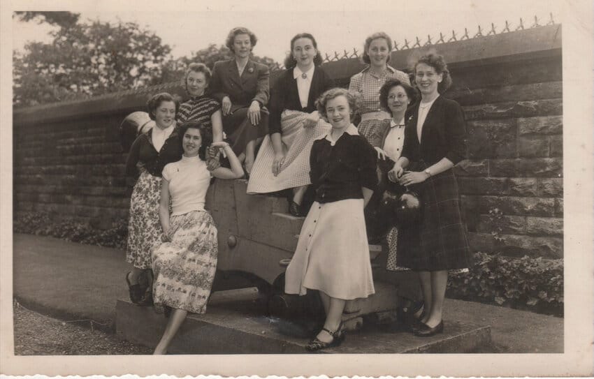 Vintage black-and-white photograph of nine young women posed together in front of a low stone wall topped with iron spikes, All are smiling at the camera. 