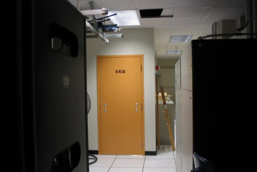 An orange door with no handle labeled '641A' at the end of a narrow hallway, flanked by tall equipment racks. A stepladder and cabinets are visible through an adjacent open doorway.