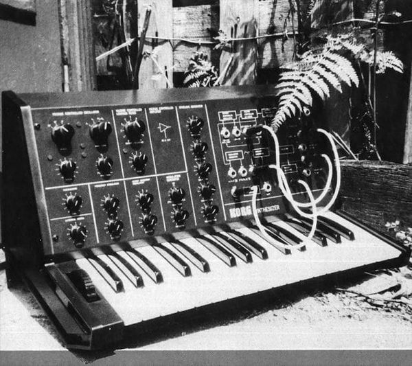 A black and white photograph of a Korg MS-10 analog synthesizer, a compact knob-covered synth with a small keyboard and patch cables, sitting outdoorswith a fern frond draped over one corner.