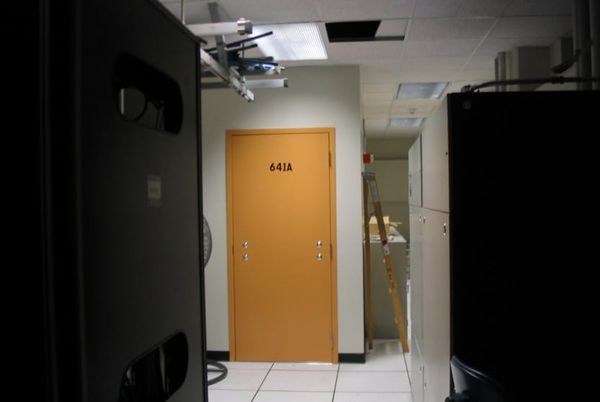 An orange door with no handle labeled '641A' at the end of a narrow hallway, flanked by tall equipment racks. A stepladder and cabinets are visible through an adjacent open doorway.