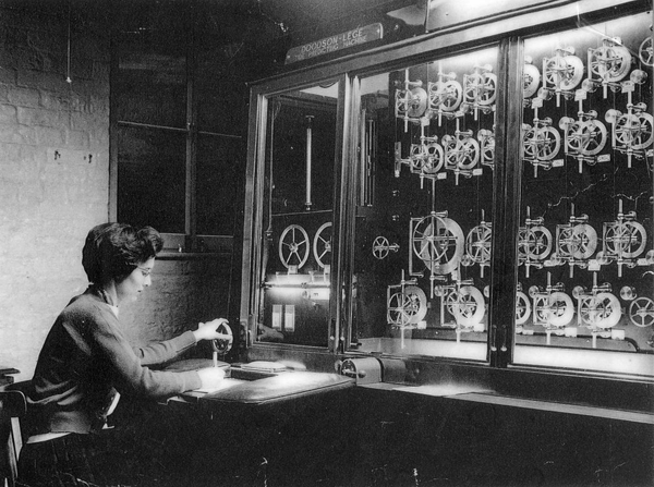 Vintage photo of a woman operating a hand crank on the Doodson-Légé Tide Predicting Machine, a large glass-fronted cabinet filled with mechanical pulleys and wheels.