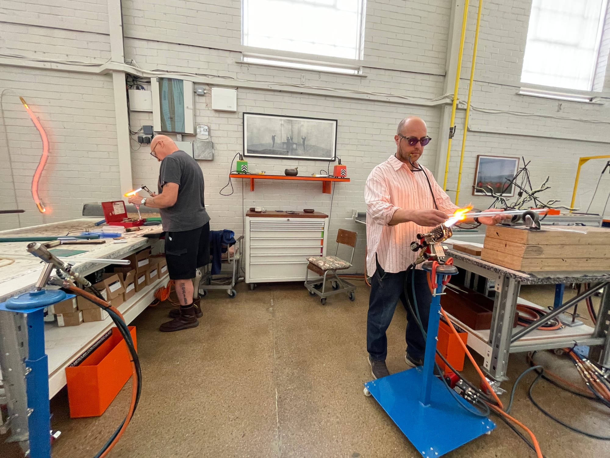Richard and Chris, standing with workbenches nearby, using small flame torches to bend glass tubing. 