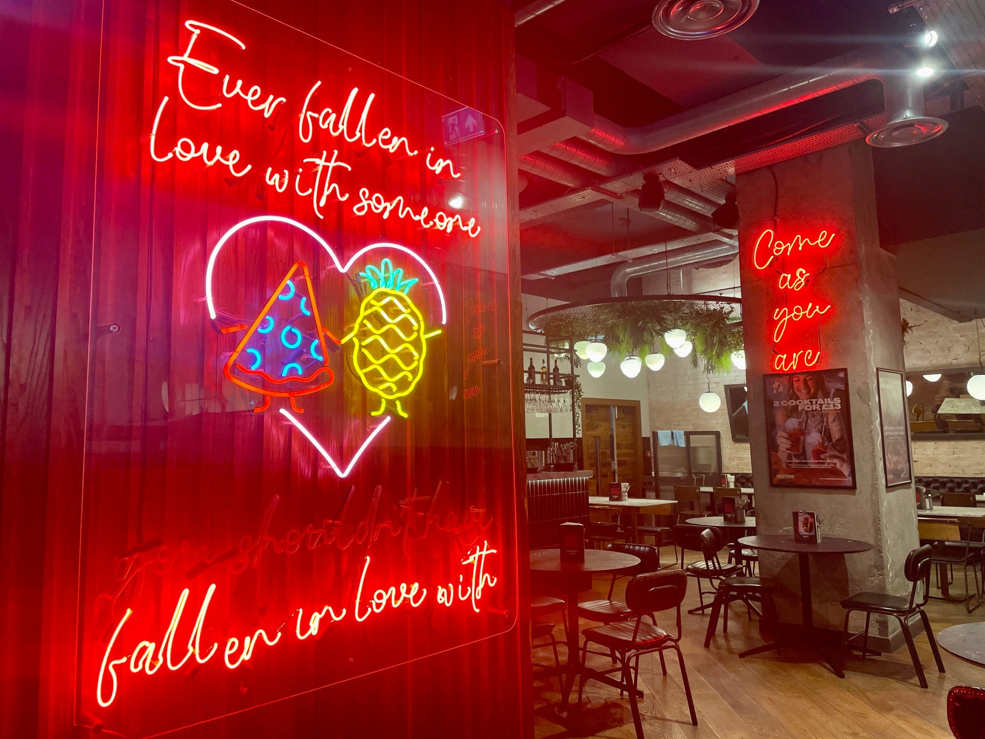 A real neon sign in a Leeds restaurant, with a more intense glow, saying "ever fallen in love with someone" showing a slice of pizza and a pineapple holding hands.