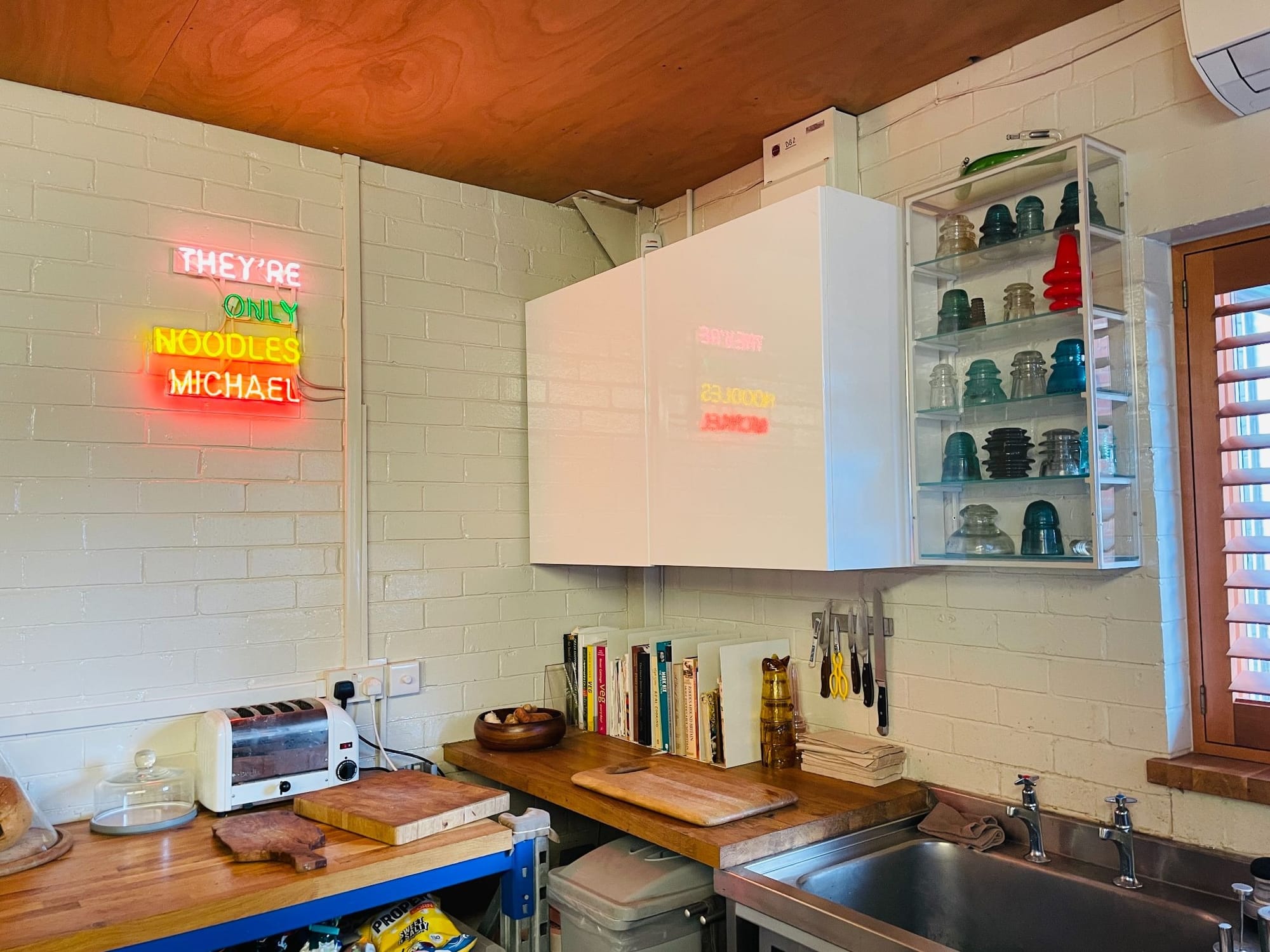 A kitchen area decorated with a neon sign saying "They're only noodles Michael" and a shelf of glass electric paraphernalia above the sink.