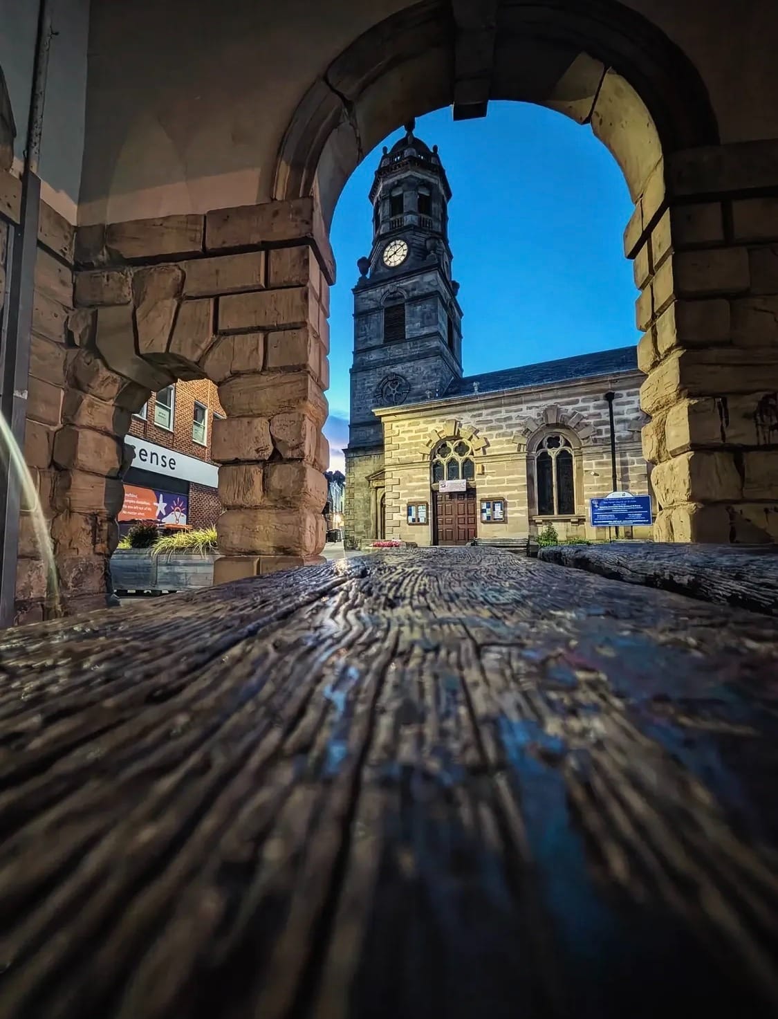 A view of the ornate tower of St Giles' Church, Pontefract, framed by the arches of the Buttercross, evening time. Much of the foreground is occupied by the textured wood of a bench.