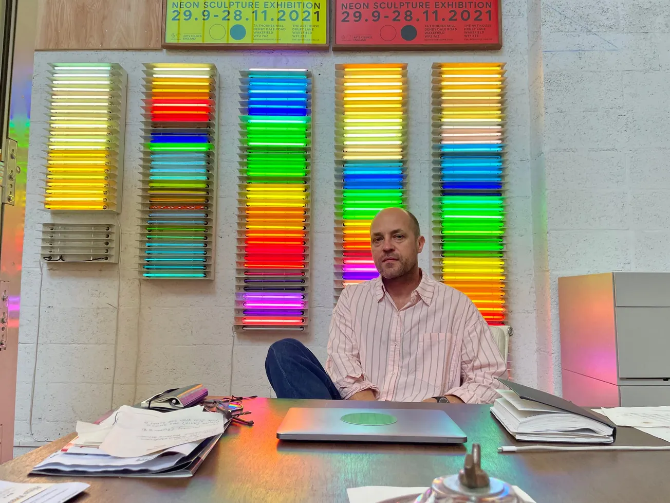 Richard William Wheater sits at his desk in front of an array of glowing neon tubing samples. 