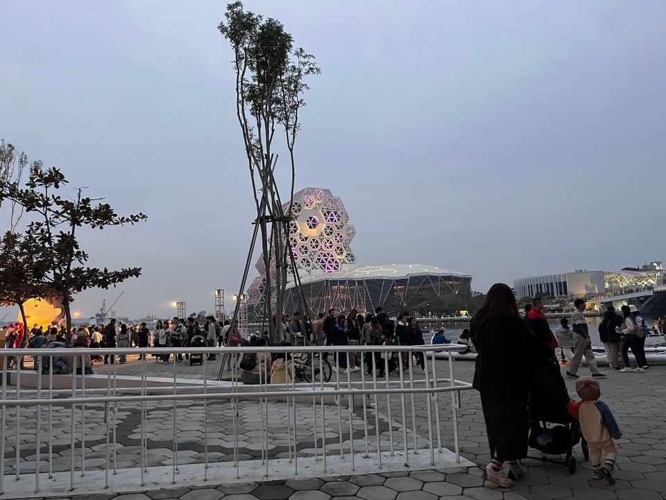 Hi-Ing Music Hall's hexagonal facade glows purple at dusk, with crowds gathering on the waterfront plaza.