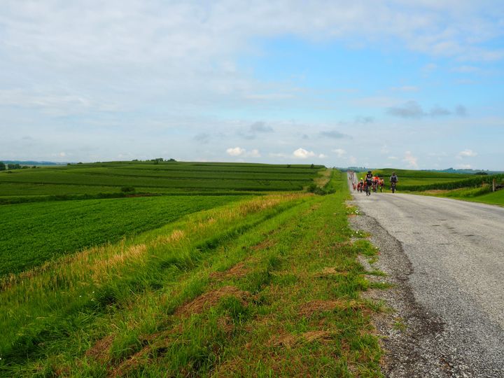 Cyclists coming up a hill on the right; lots of crop fields on the left with open skies all around.