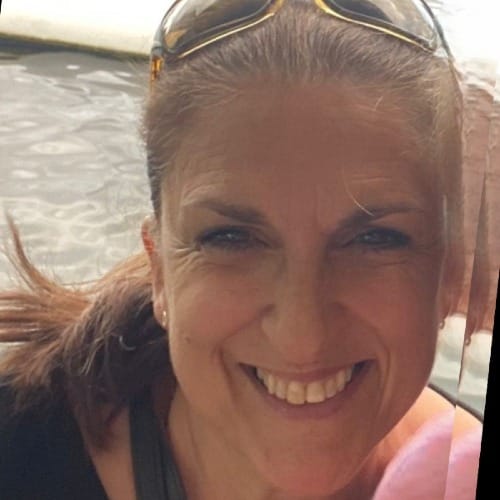 Smiling woman with glasses propped up and over her light brown hair with water scene behind her.