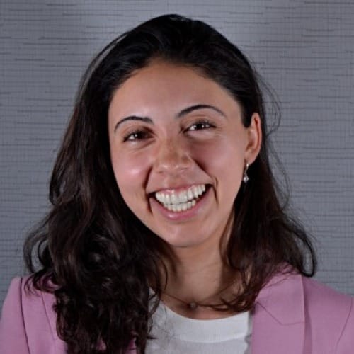 Young woman with dark hair below shoulders brushed to one side smiling and wearing pink jacket and white shirt