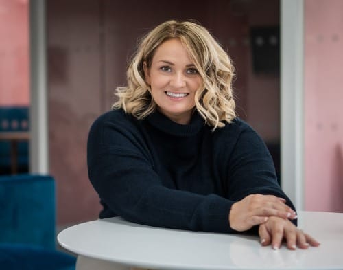Blonde woman with shoulder length hair emiling with thick dark blue sweater and hands clasped before her on white table