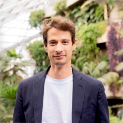 Man with short brown hair blown slightly upwards with white t shirt and dark jacket and green forestation in background