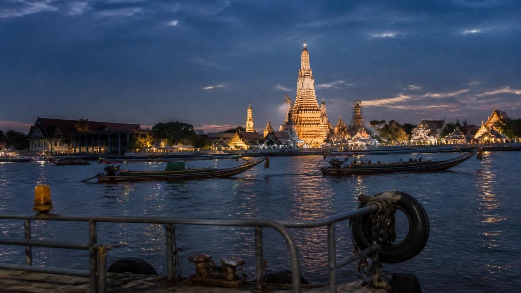 Long-tail boats crossing the Chao Phraya River at blue hour, with the spires of Wat Arun lit on the far bank.