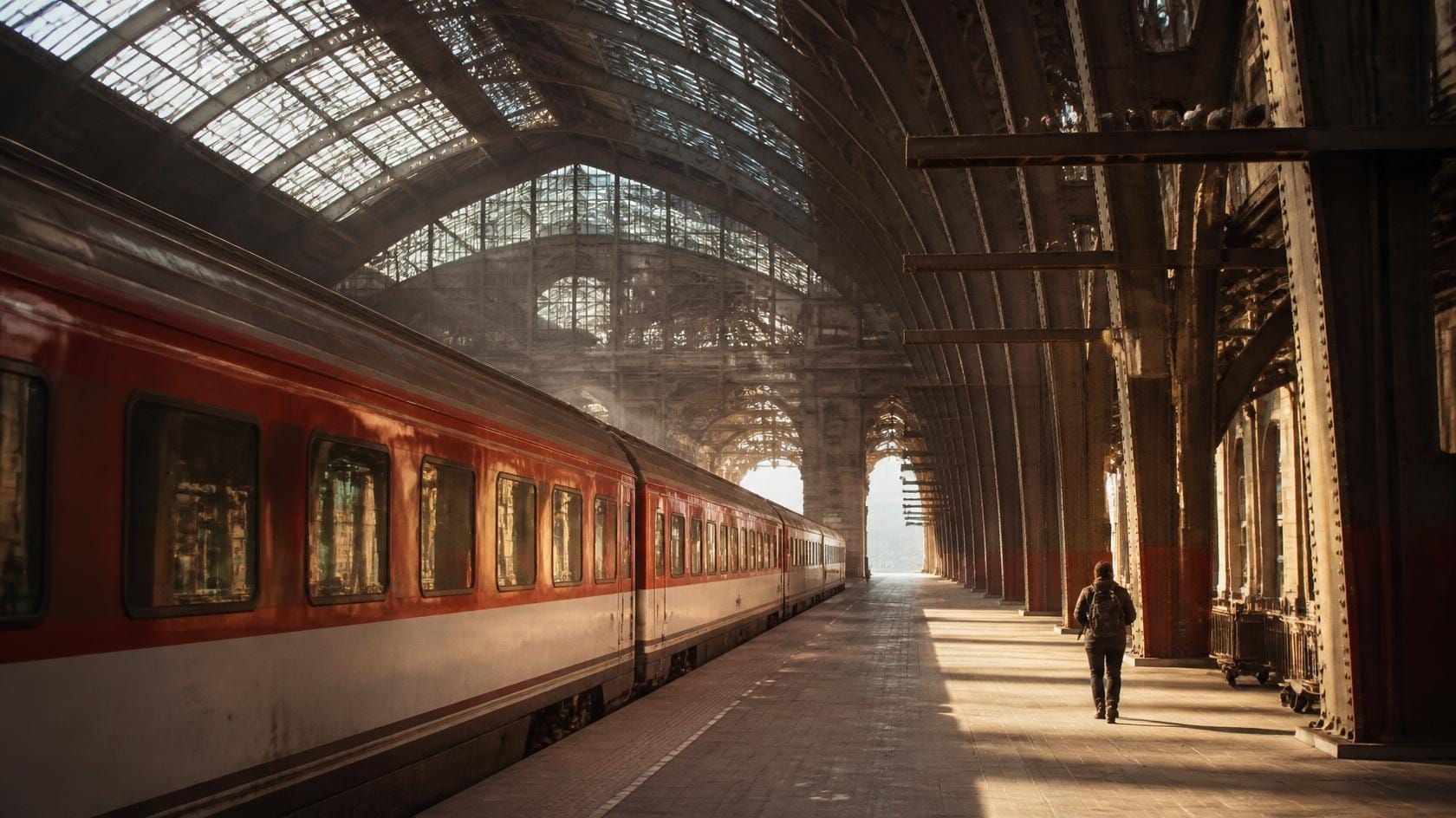 A traveller with a backpack walks along a sunlit platform under the iron arches of a grand European railway station towards a red train.