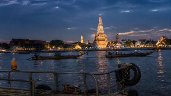 Long-tail boats crossing the Chao Phraya River at blue hour, with the spires of Wat Arun lit on the far bank.