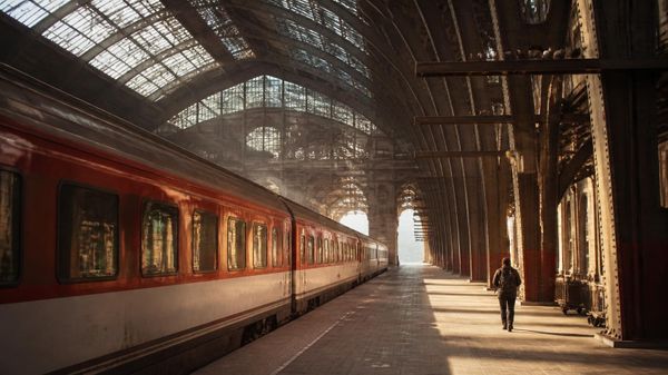 A traveller with a backpack walks along a sunlit platform under the iron arches of a grand European railway station towards a red train.