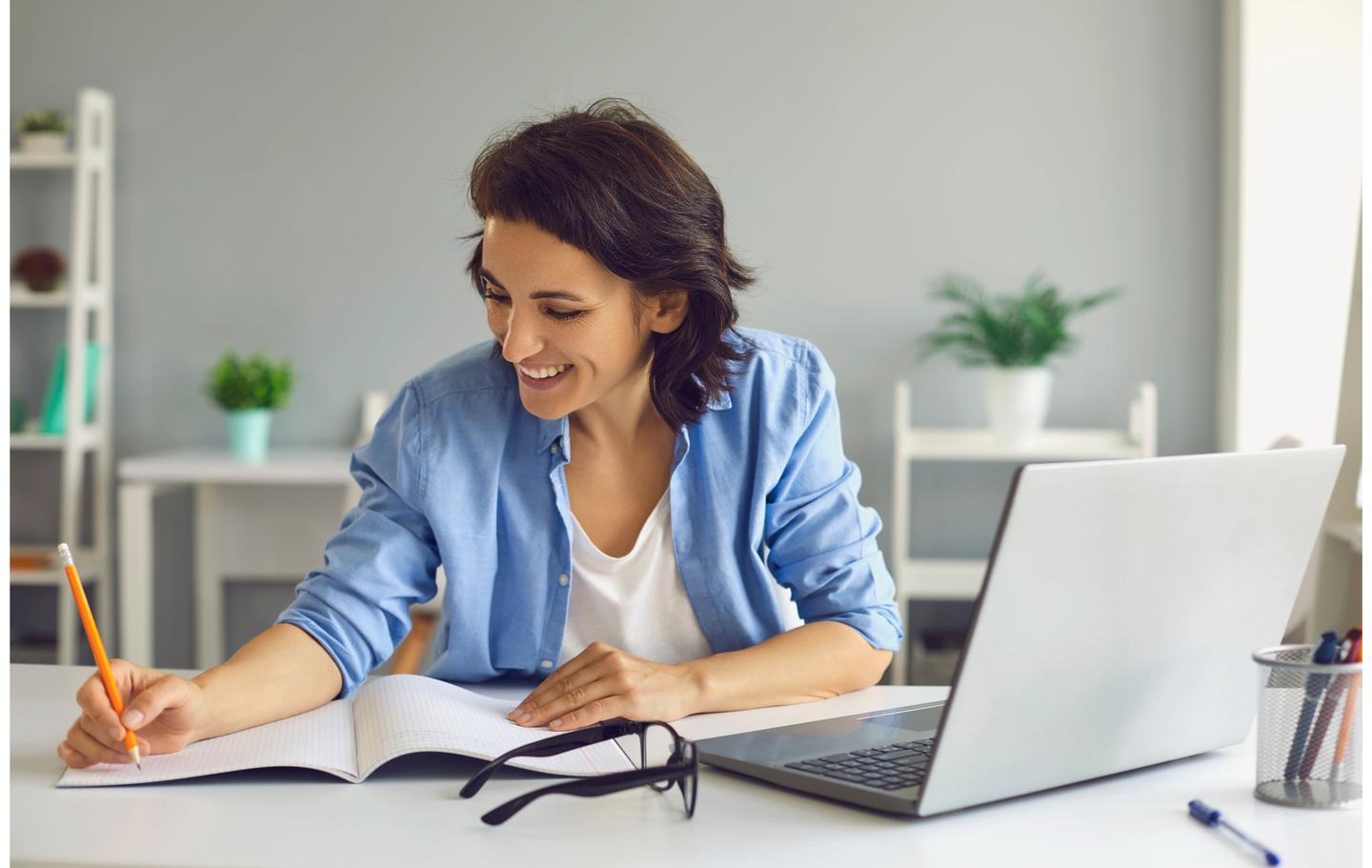 Smiling woman sitting at a desk, writing in a notebook while working on a laptop, in a bright home office setting
