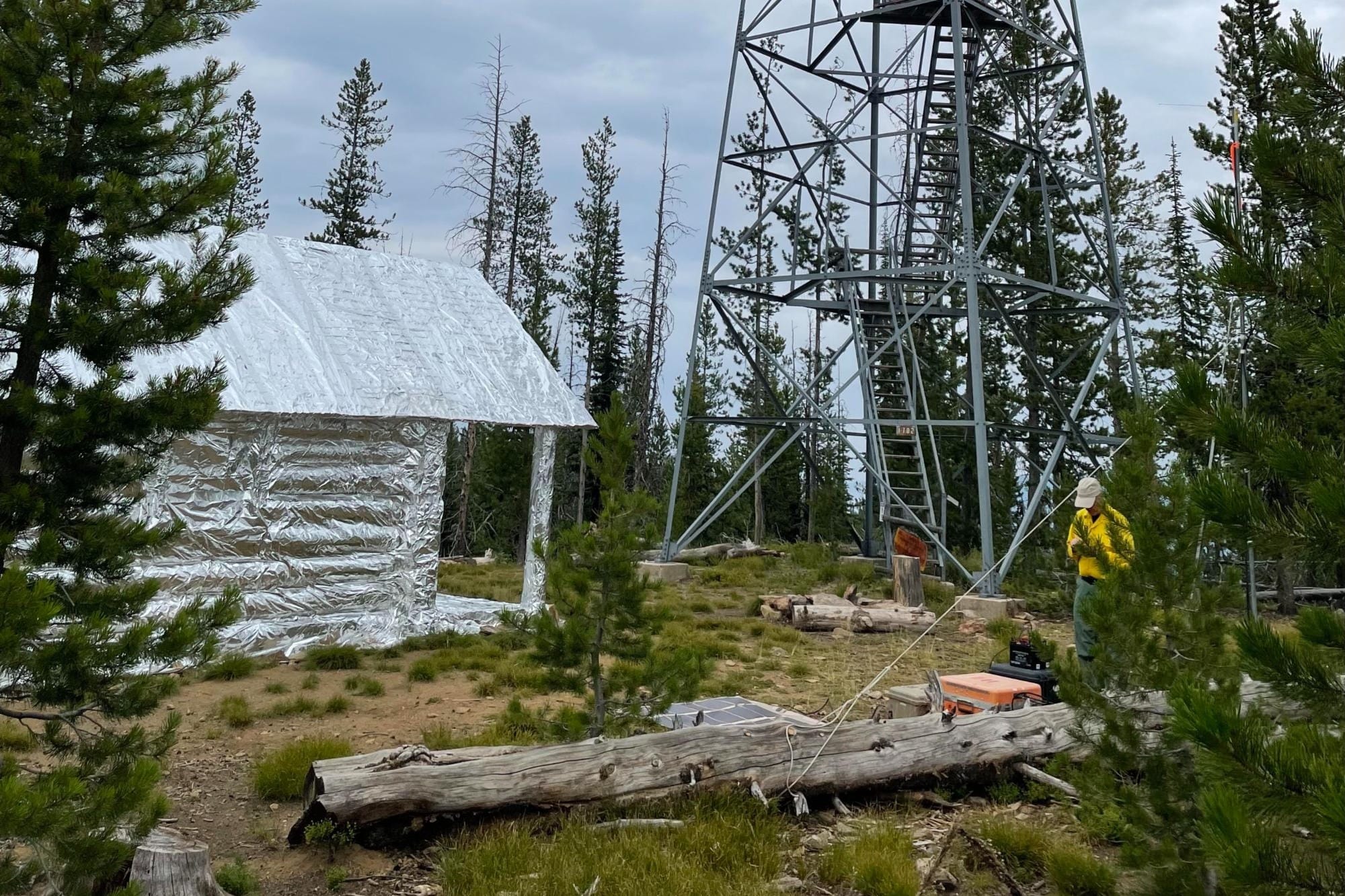 A small cabin fully covered in silver structure wrap beside the steel tower of Arctic Point Lookout, with a firefighter in a yellow shirt nearby