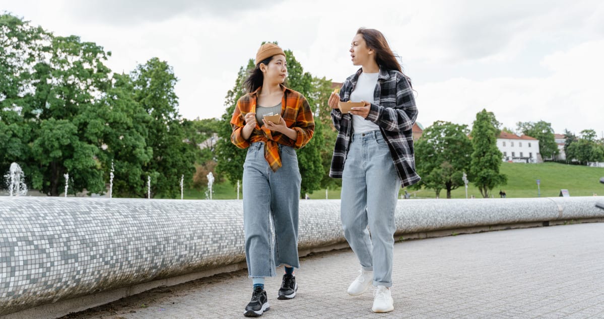 Two women enjoying a casual walk in the park, chatting and eating snacks outdoors.