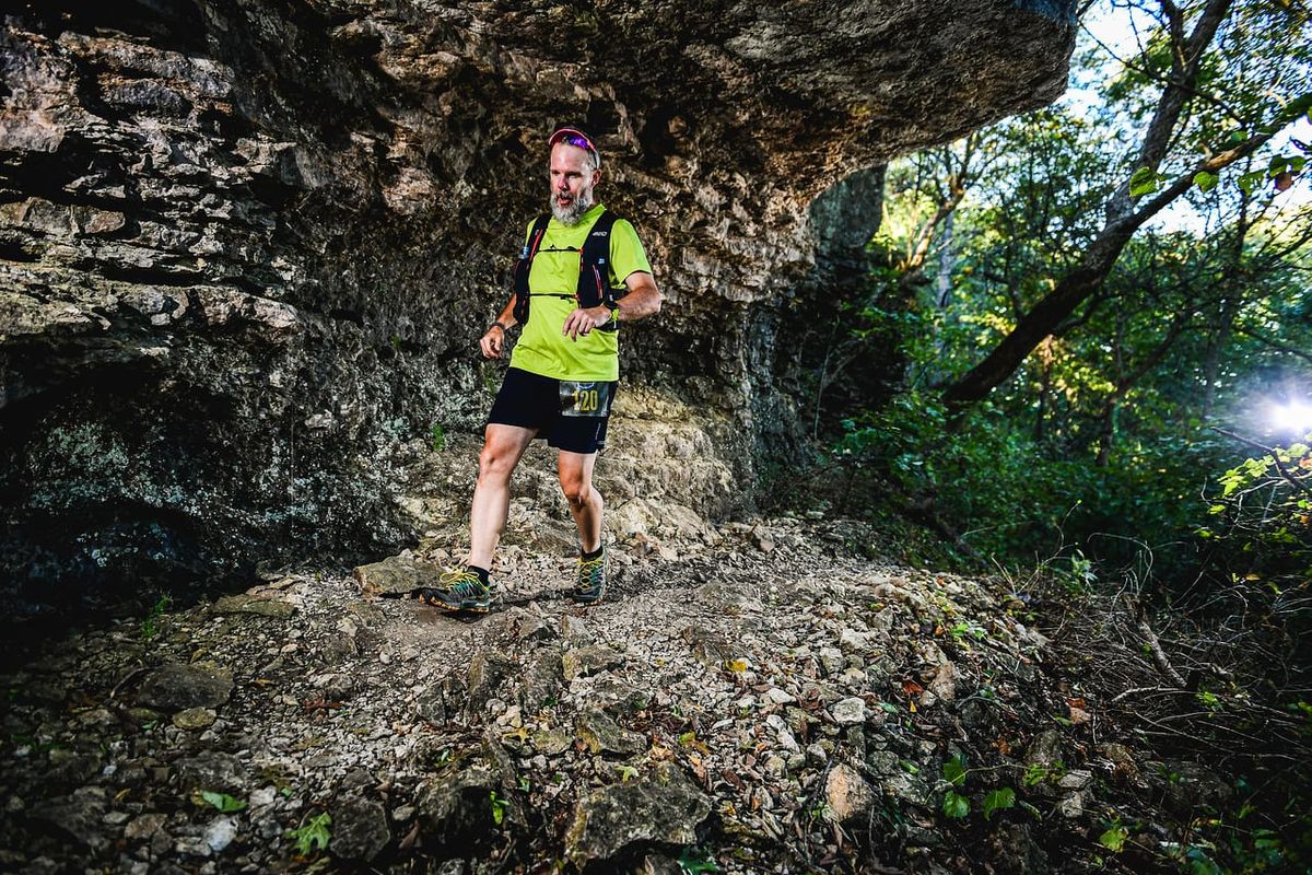 Matt running over a very rocky trail next to a stone bluff with trees in the background.