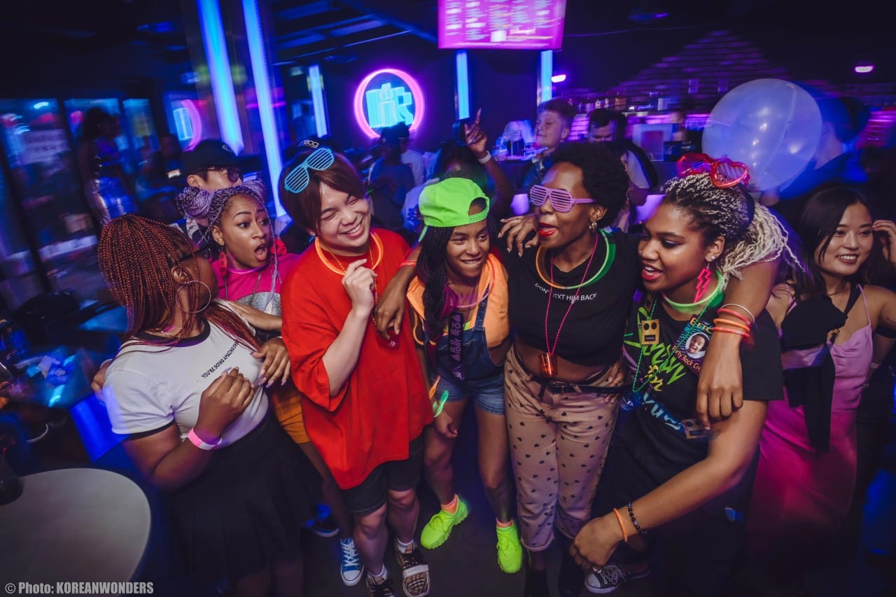 Diverse crowd dancing at Lit Lounge Itaewon, wearing neon outfits and glow sticks during a weekend nightlife party in Seoul