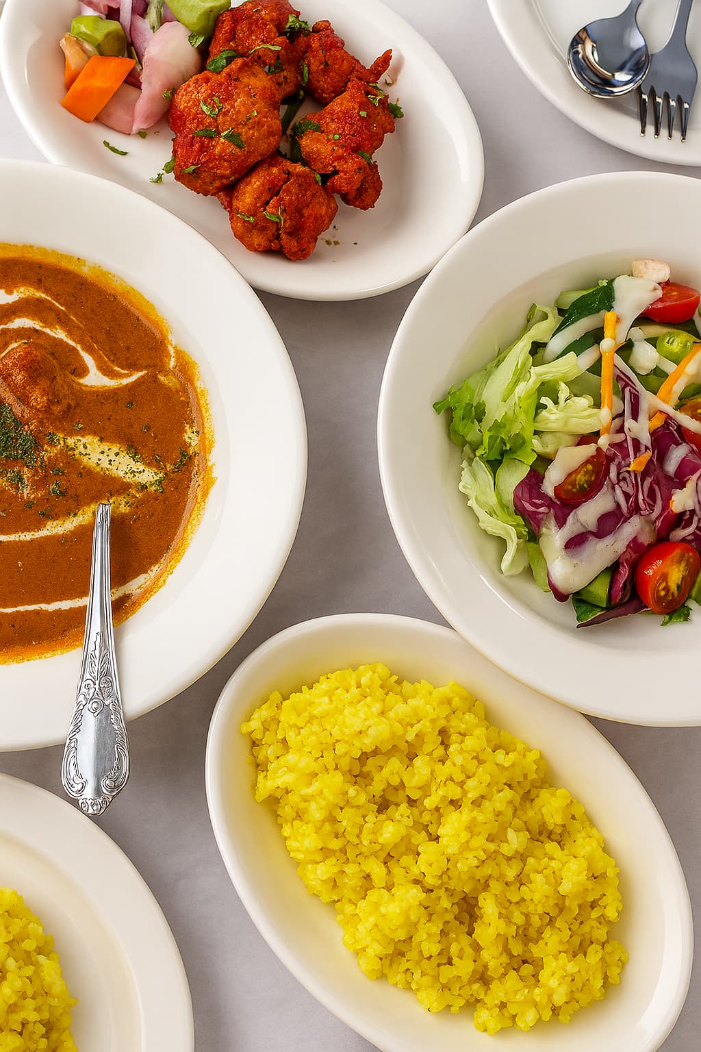 A vibrant Indian lunch set featuring curry with cream swirl, yellow turmeric rice, fresh salad with dressing, and tandoori chicken served on white plates.