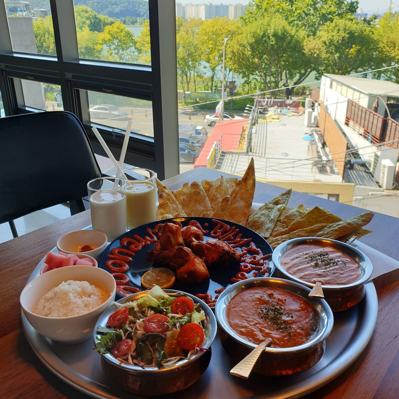 Indian lunch set at Poonawala Indian Restaurant with curry, naan, rice, salad, tandoori chicken, and lassi, served by a window with a city and river view.