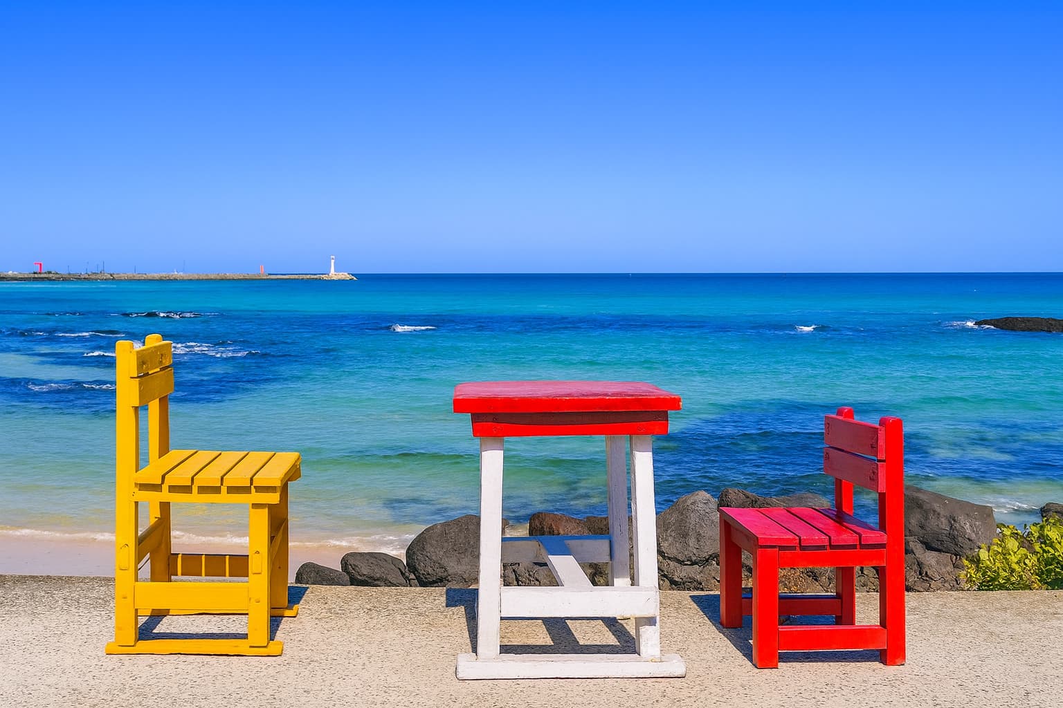 Colorful red and yellow chairs with a small table facing the turquoise ocean waves at a Jeju beach under a clear blue sky
