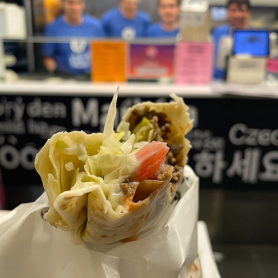 Close-up of a half-eaten kebab wrap with lettuce, tomato, and beef in Daegu restaurant with staff in blue uniforms blurred in background