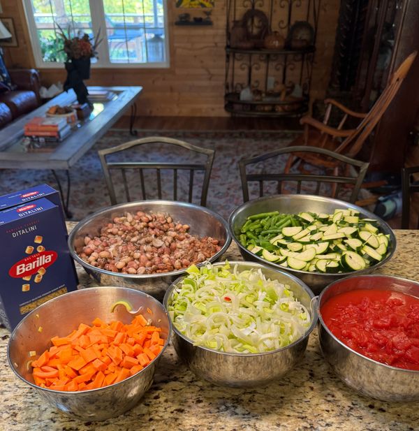 Silver mixing bowls filled with colorful vegetables that have been sliced, diced, minced, etc. sit in a countertop with a bowl of cannoli beans and box of pasta. 
