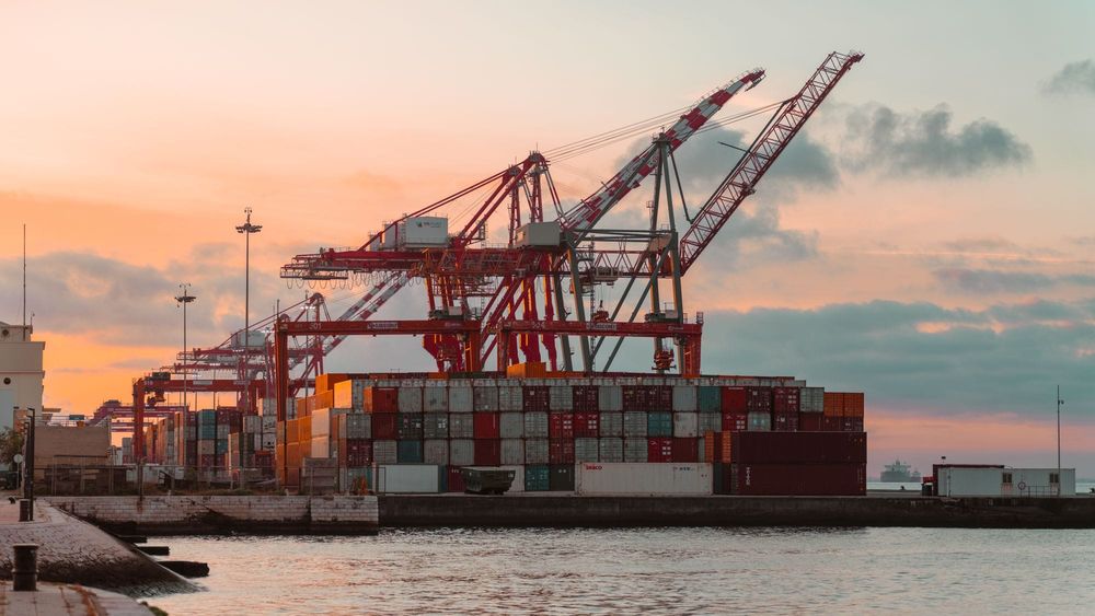 Red container cranes over stacked shipping containers at a waterfront port under a pink and orange sunset sky
