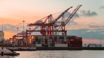 Red container cranes over stacked shipping containers at a waterfront port under a pink and orange sunset sky