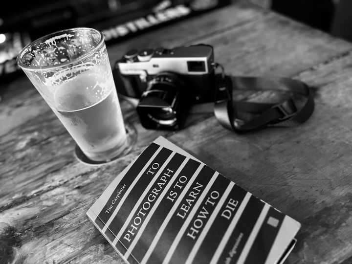 A copy of the book To Photograph is to Learn How to Die on a wooden bar next to a camera and a pint glass of beer