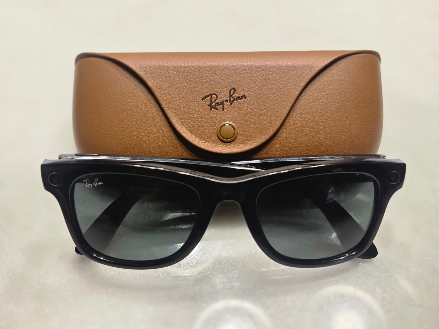 A pair of shiny black Ray-Ban Meta Gen 1 smart glasses positioned in front of their brown leather charging case.