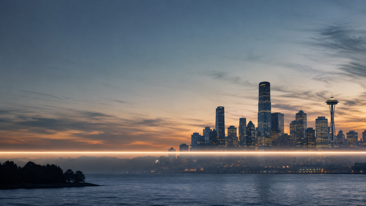 Seattle skyline at dusk across Puget Sound, with a bright horizontal band of light cutting level through the frame below the towers.