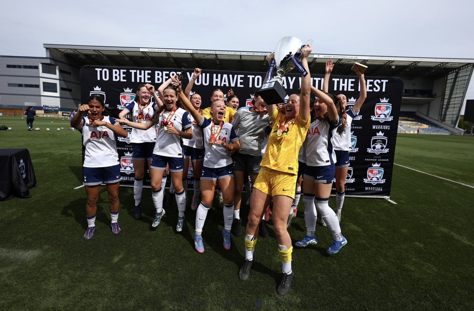 The Women's U16s lift the PGA trophy in front of a banner that says, "To be the best you have to play with the best."