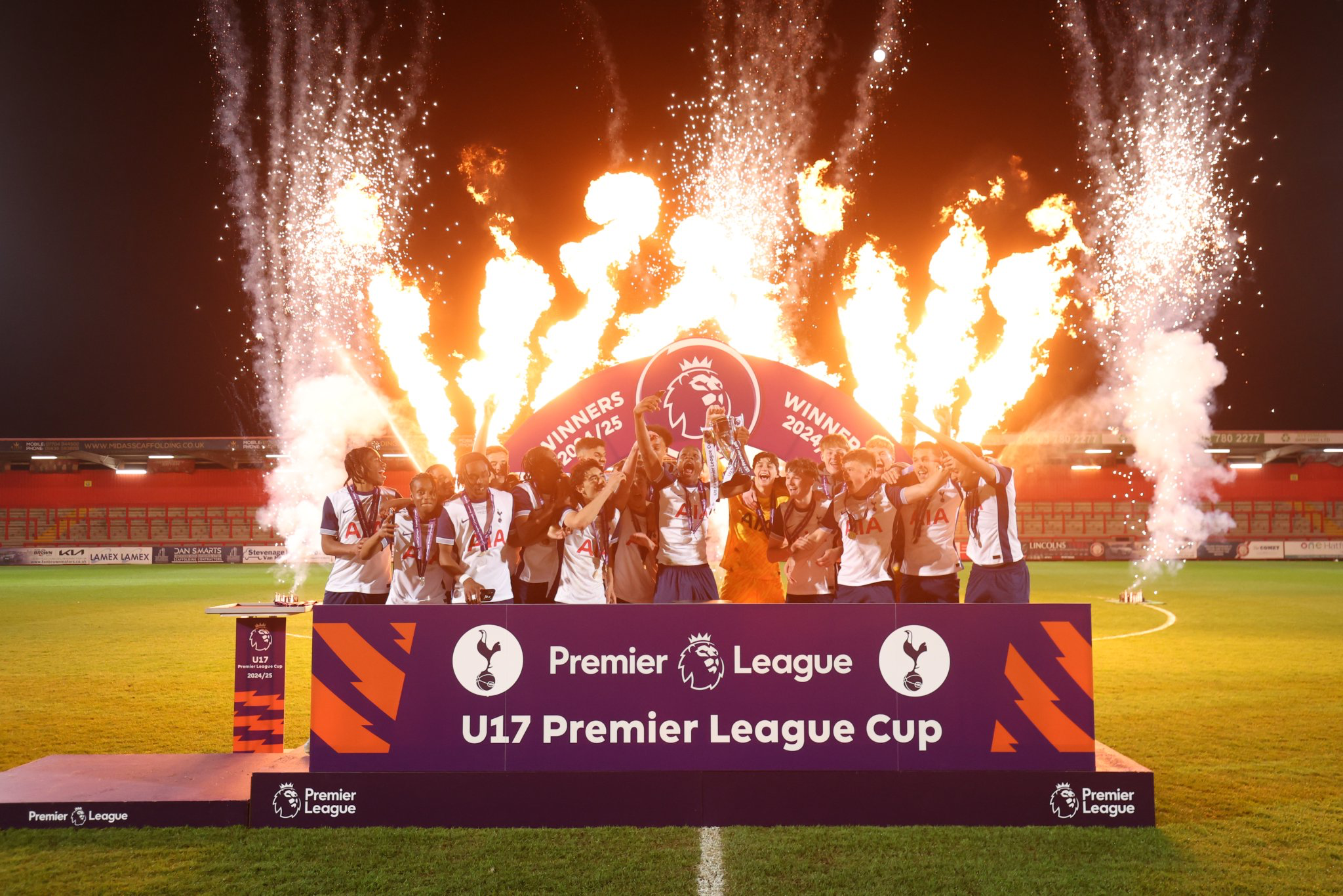 The Men's Academy U17s lift the Premier League Cup trophy with fireworks and flares in the background.