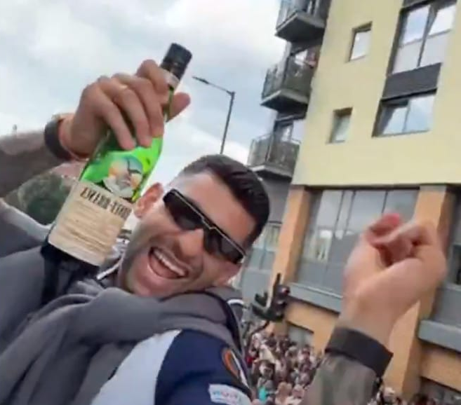 Cuti Romero holds up a bottle of fernet branca with shades on and his hoodie tied around his neck, his other hand raised in a fist, a goofy smile on his face.