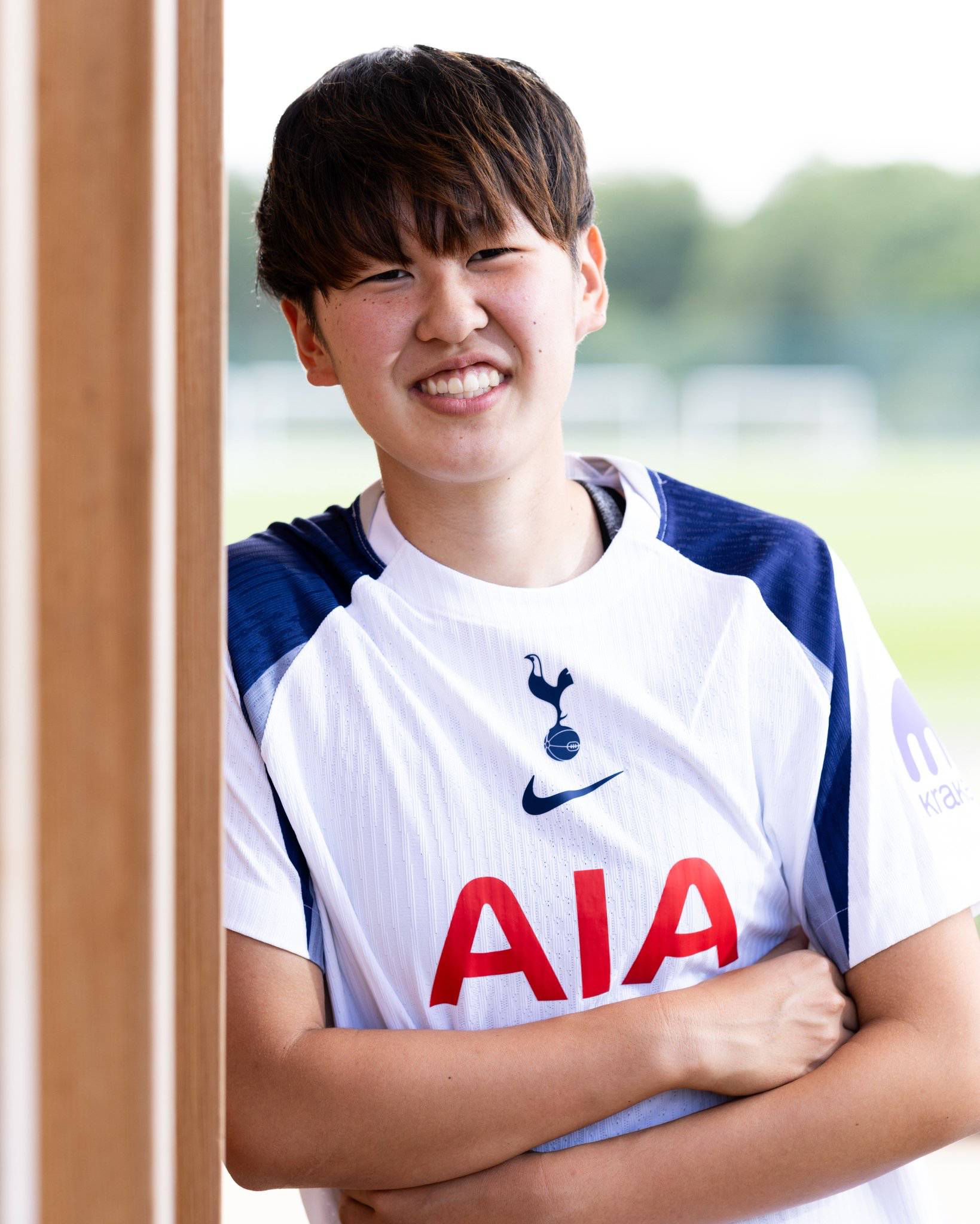 Tōko Koga leans against a wall with her arms crossed, in her new Spurs home kit, smiling broadly.